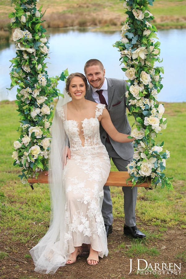 The bride and groom are sitting on a swing covered in flowers for their wedding.