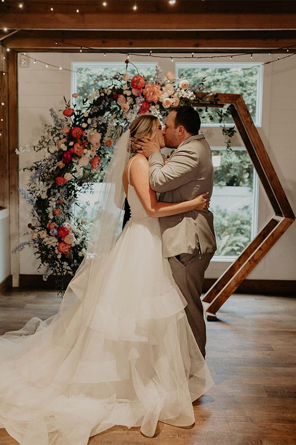 The bride and groom kissing with flowers in the back.
