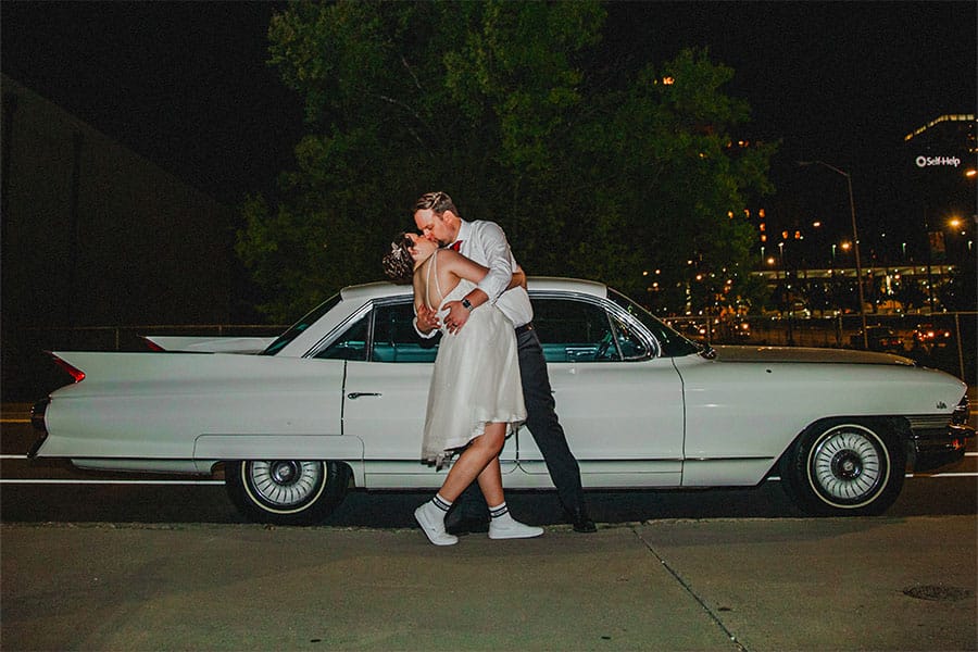 The bride and groom kissing in front of a car.